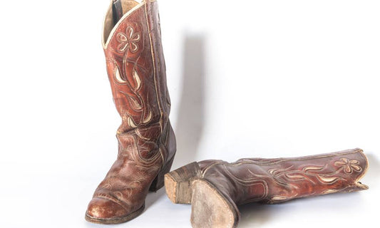 A worn pair of ornate brown cowboy boots with one boot laying on its side in front of a white background.