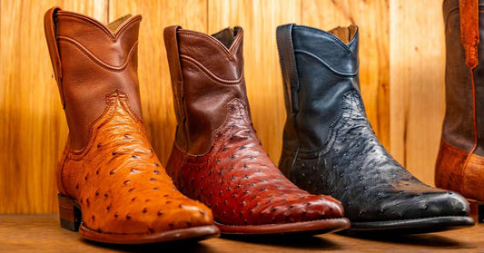 Three exotic leather western boots made from tan, red, and black ostrich leather sit on a wooden shelf for display.