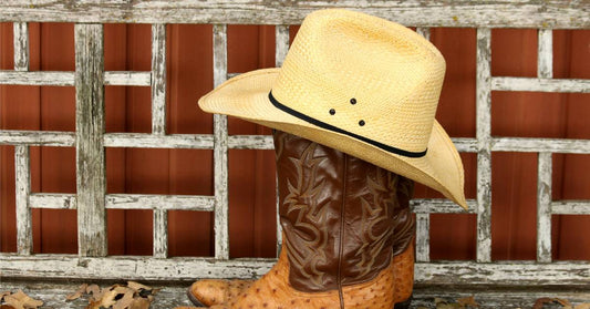 A straw cowboy hat sits on top of a pair of ostrich leather cowboy boots. The boots sit on a weathered porch.