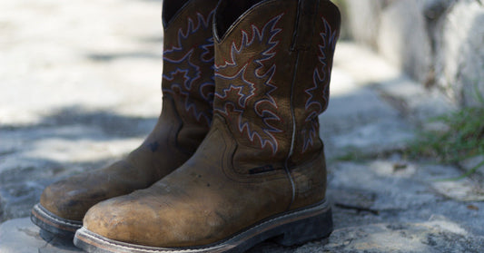 A pair of brown pull-on Western work boots sit in the shade on the concrete outside with the sun casting a soft shadow.