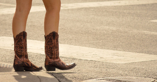 A woman walks across a concrete crosswalk wearing brown mid-calf boots with detailed embroidery on the shaft.