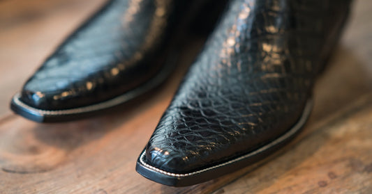 A pair of handmade black exotic leather boots with white stitching and a snip-toe sit on a weathered wooden floor.