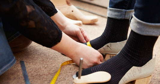 A shoemaker measuring a person's feet that are resting on wooden heels, with other shoemaker tools are scattered nearby.