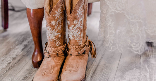 A light-brown pair of embroidered boots sits on a wooden floor in front of a chair with a white lace dress in the background.