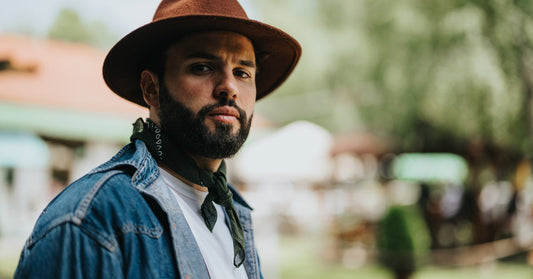 A bearded man wearing a hat, white shirt, and denim jacket stands outdoors with a furrowed brow, looking serious.